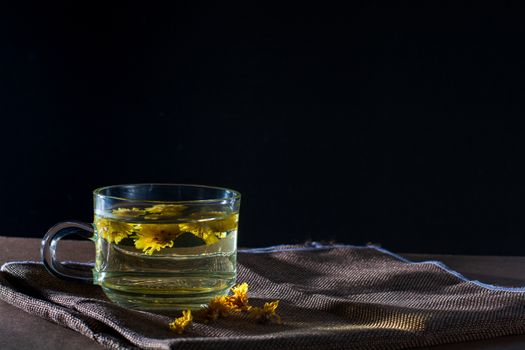 Cup of Chrysanthemum tea with dry flower on brown table cloth on black background. Healthy beverage for drink. Herbs and medical concept.  