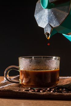 Pour hot of black coffee from green moka pot to clear coffee cup with smoke and coffee beans on brown table cloth and wooden table. Benefit of coffee concept.