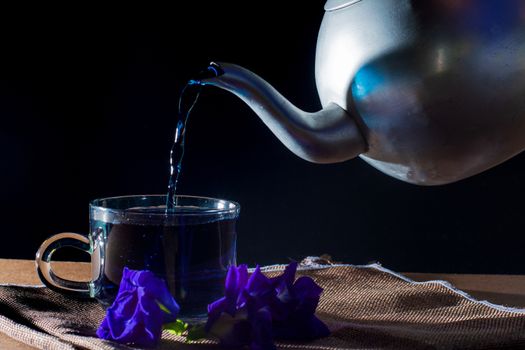 Pour Butterfly pea tea by metal pot to cup with violet flower on brown table cloth on black background. Healthy beverage for drink. Herbs and medical concept. 

