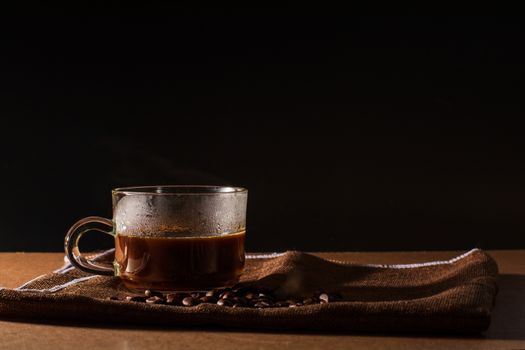 Cup of hot coffee with smoke and group of coffee beans on brown table cloth with black background with copy space for your text. Benefits of coffee concept.