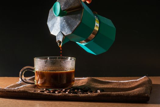 Pour hot of black coffee from green moka pot to clear coffee cup with smoke and coffee beans on brown table cloth and wooden table. Benefit of coffee concept.