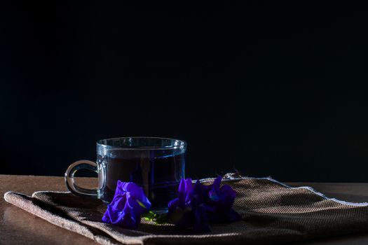 Cup of Butterfly pea tea with fresh violet flower on brown tablecloth and wooden table on black background. Healthy beverage for drink. Herbs and medical concept.