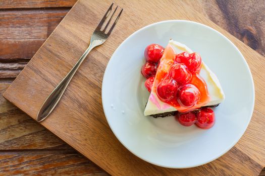 Cherry cheese pie cake in white plate and metal fork on wooden table.