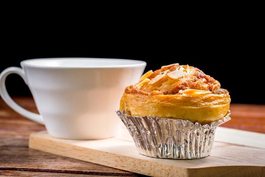 Delicious homemade almond danish bread in foil cup and white cup of coffee on a wooden chopping board and table in the home kitchen on black background.