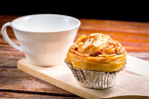 Delicious homemade almond danish bread in foil cup and white cup of coffee on a wooden chopping board and table in the home kitchen on black background.