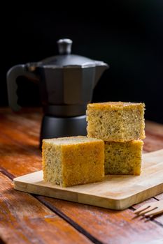 Close up square cut of homemade sweet and solf banana cake on wooden chopping board and fork on table with solf focus black moka pot. Delicious and healthy bakery.