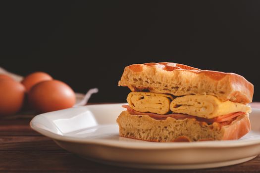 Waffle sandwich with egg and chicken ham in white dish on wooden table and black background. Homemade food concept.