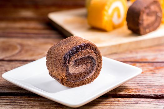 Close up delicious chocolate roll cake with whipping cream in a square white plate on a wooden table at the home kitchen with a black background and copy space. Homemade bakery concept.