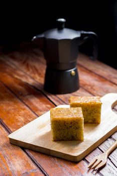 Close up square cut of homemade sweet and solf banana cake on wooden chopping board and fork on table with solf focus black moka pot. Delicious and healthy bakery.