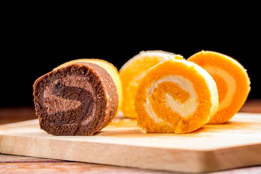 Close up delicious chocolate, vanilla and orange roll cake with whipping cream on  a wooden chopping board and table at the home kitchen with a black background and copy space. Homemade bakery concept.