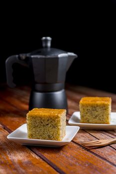 Close up square cut of homemade sweet and solf banana cake on white plate on table with solf focus black moka pot. Delicious and healthy bakery.