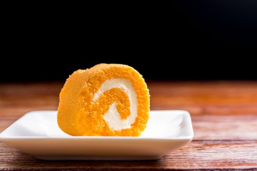 Close up delicious orange roll cake with whipping cream in a square white plate on a wooden table at the home kitchen with a black background and copy space. Homemade bakery concept.