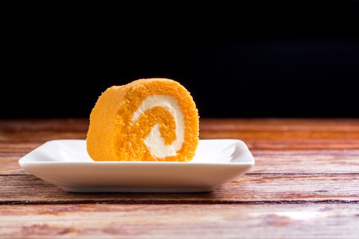Close up delicious orange roll cake with whipping cream in a square white plate on a wooden table at the home kitchen with a black background and copy space. Homemade bakery concept.
