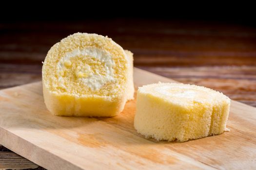 Close up delicious vanilla roll cake with whipping cream in a square white plate on a wooden table at the home kitchen with a black background and copy space. Homemade bakery concept.