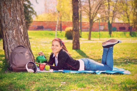 Beautiful smiling young girl lying on the green grass in the park, inserting a coin in a green piggy bank. Savings and money concept. Student saving concept.