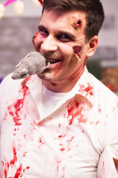 Man dressed up like a bloody zombie with a rat in his mouth at halloween celebration.