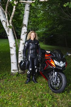twenty something blond woman, wearing full motocycle gear, standing beside sport motocyle, in a park