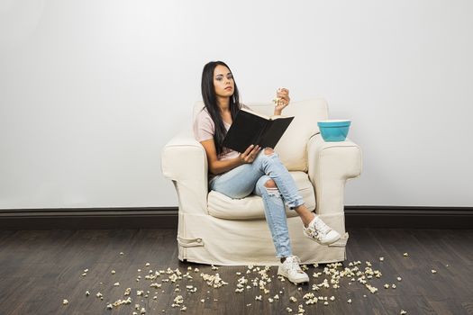 young woman, making a mess of popcorn around a white couch, while reading a hard cover book