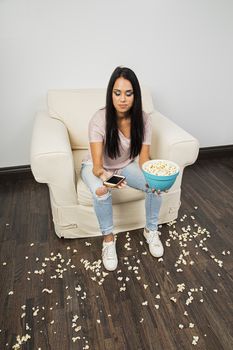 young woman, sitting on a white couch, texting on her smart phone, eating popcorn and making a mess