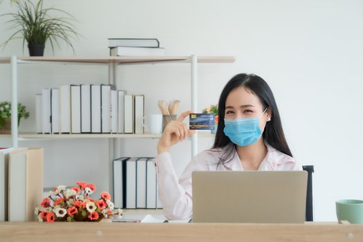 A beautiful Asian woman is working at home happily. She is using a laptop to work. She wore a mask to prevent the outbreak of the virus. She held a credit card in her hand.( Mock up  credit card )