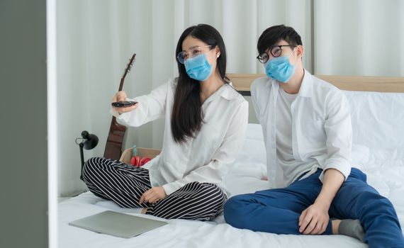 Social distancing.Young Asian people are watching television news. Stay in the bedroom happily. They wear masks to prevent the outbreak of the virus.
