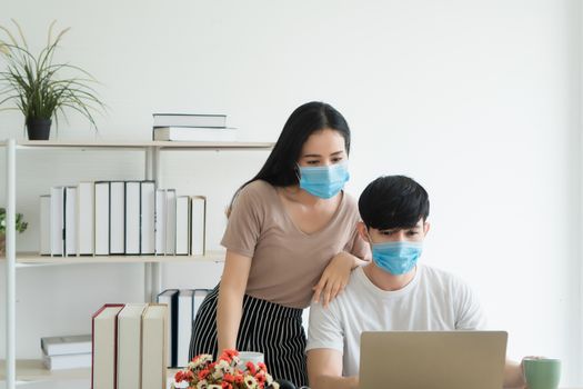 Working from home - Concept. Asian young people are working together at home happily. They are using laptops for work. They wear masks to prevent the spread of the virus.