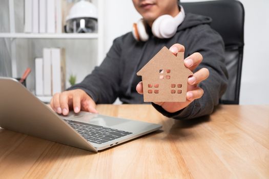 Smiling businessman holding a wooden house in home workspace. The concept of working from home until the end of Coronavirus (COVID-19) quarantine, happily freelance working lifestyle