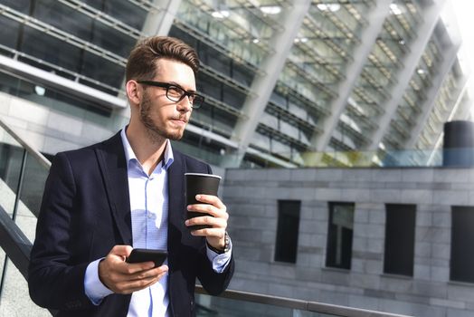 Handsome successful businessman in elegant suit outdoors stand at street in the city with coffee.
