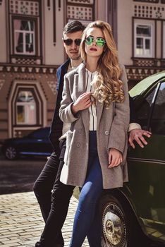 Fashionable couple in sunglasses stand at street near the car