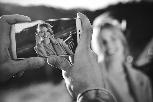 A man make a portrait on the phone of happy smiling woman standing on the bridge on sunny spring or autumn day outdoor