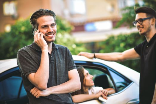 Handsome young caucasian man talking on the mobile phone and smiling while standing near the car with his friends chatting in the background.
