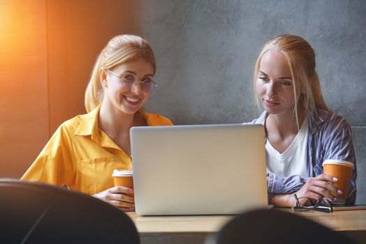 Two cute girls in casual clothes are looking at the screen of a laptop while sitting in office. Bloggers or designers are reading news on a laptop while having coffee break.