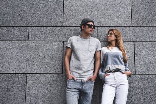 Two friends leaning to a wall and ignoring each other. Technology addiction concept, guy and a woman in gray wall background.