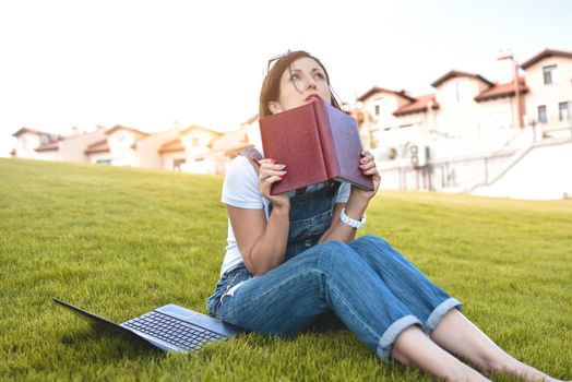 Portrait of pretty young woman sitting on green grass in park with laptop on legs, spending summer day working outdoor, using laptop and wireless Internet for online work. Lifestyle concept.