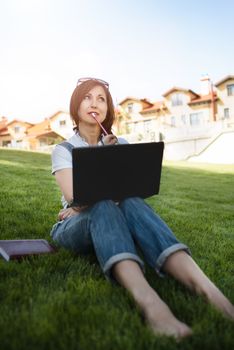 Portrait of pretty adult woman sitting on green grass in park with laptop on legs, spending summer day working outdoor, using laptop and wireless Internet for online work. Lifestyle