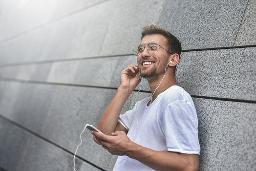 Earphones concept. People, technology, travel and tourism - man with earphones, smartphone on city street and listening to music over gray wall background