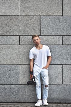 Picture of handsome Caucasian guy on grey textured wall in white T-shirt and white jeans with camera hanging on strap on his arm.