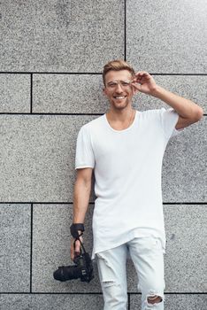 Picture of handsome Caucasian guy on grey textured wall in white T-shirt and white jeans with camera hanging on strap on his arm.