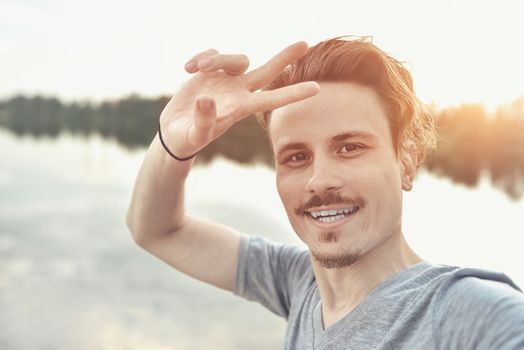 portrait of Young handsome stylish smiling guy makes selfie against the lake. beautiful nature