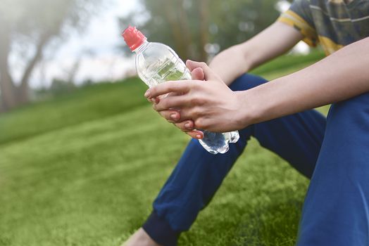 Young caucasian man runner relaxing holding drinking water bottle and sitting on grass in the park outdoors after sport at early morning time, Exercise and healthy concept. summer time