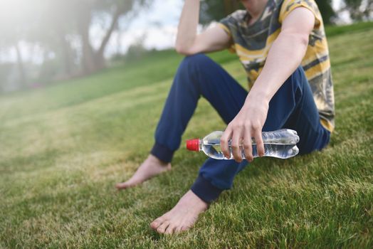 Young caucasian man runner relaxing holding drinking water bottle and sitting on grass in the park outdoors after sport at early morning time, Exercise and healthy concept. summer time