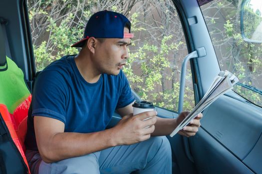 A young Asian man who is passionate about reading He is reading a newspaper at the car. He is a truck driver. He uses his free time to read books and to learn more.