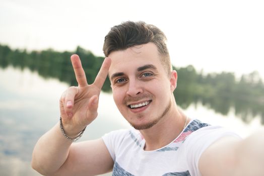 portrait of Young handsome stylish smiling guy makes selfie against the lake. beautiful nature