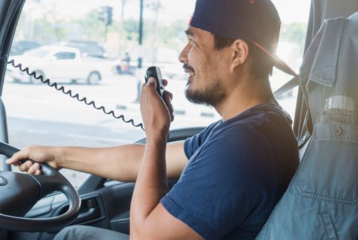 An Asian male truck driver is driving down the road with caution. He is using radio to communicate with the call center.