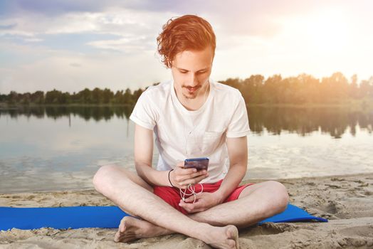 Man is sitting on the beach at lake and writing a sms on phone. good mood. beautiful view