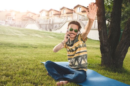 young stylish handsome man is sitting on grass, resting and talking on the phone. waves his hand welcoming friends