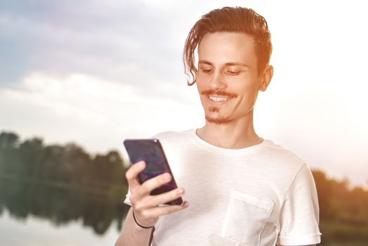 Young man walking near beautiful bay using phone and headphones for communicate with friends . good mood, beautiful view