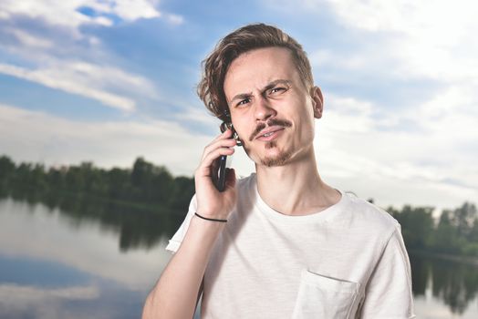 Portrait of angry confused aggressive in bad mood guy irritably speaks on the phone on the background of the lake