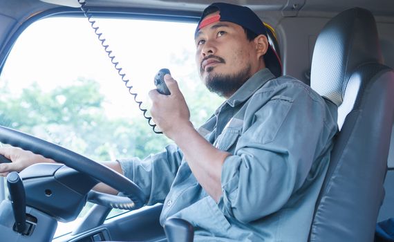 An Asian male truck driver is driving down the road with caution. He is using radio to communicate with the call center.