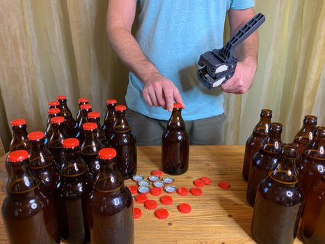 Craft beer brewing at home, man closes brown glass beer bottles with plastic capper on wooden table with red crown caps. Horizontal stock image